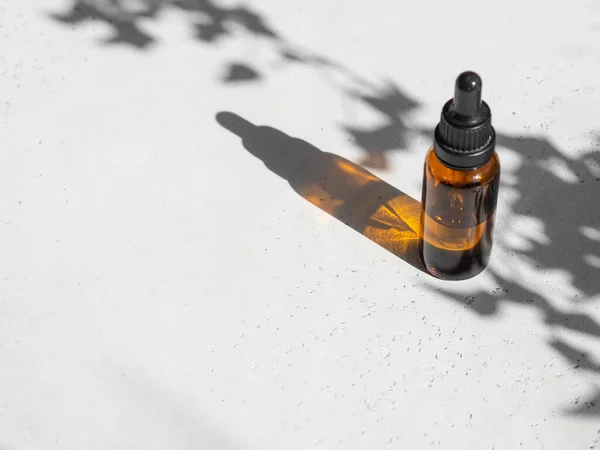 Brown glass with a dropper. Bottle mockup cosmetic oil dropper on grey background and shadows of plants. Copy space