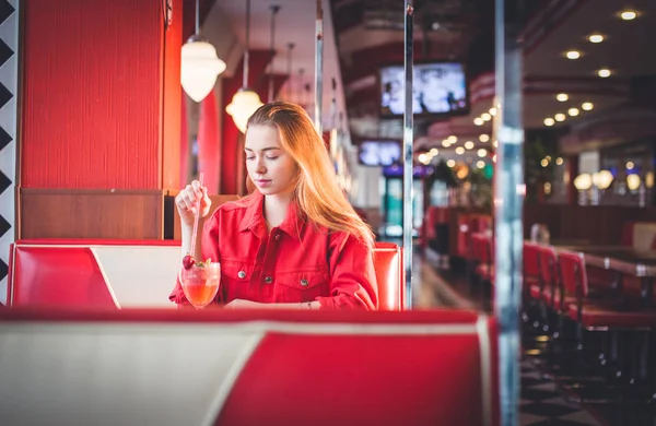 Открытый солнечный портрет стильной веселой женщины с красными губами, кудрявая блондинка, сидящая на террасе кафе в центре города, пьющая вкусный сладкий клубничный коктейль и смеющаяся . — стоковое фото