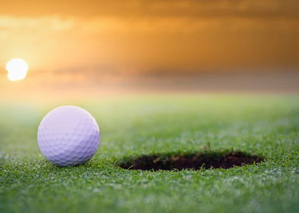 Bola de golfe sentado em um campo de golfe colocando verde ao lado do buraco. com um belo pôr do sol . — Fotografia de Stock