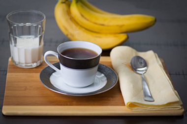 Buhar ve bir bardak süt ve muz bir kaşık ve peçete ile ahşap bir masa üzerinde arka planda bir demet ile sıcak siyah kahve.