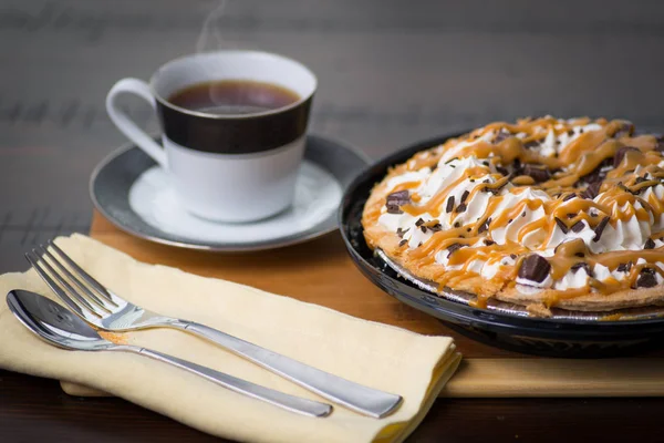 Hot fresh black coffee steam, with a caramel chocolate whip cream pie cookie crust, on a round dark brown wooden table, fork and spoon tan napkin