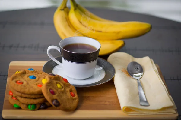 Café caliente de vapor negro, con galletas de chocolate y plátanos amarillos sobre una mesa de madera con una cuchara y servilleta bronceada . Fotos de stock libres de derechos