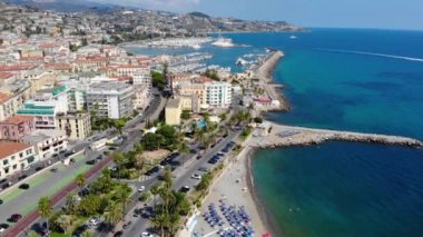 Deniz kıyısındaki güzel İtalyan şehrinin muhteşem hava manzarası. Plajların üzerinde ve palmiye ağaçlarıyla sahil boyunca uçan insansız hava aracı. San Remo, İtalya 4K