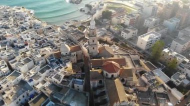 Güneşli bir günde deniz kenarında bir İtalyan şehri. Kilisenin, evlerin ve denizin havadan görünüşü. Monopoli, Puglia, Bari, İtalya 4K
