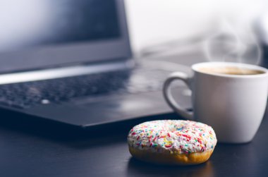 Çörekli bir fincan kahve. Arka planda bilgisayar. Kahve köpüğü. Yiyecek, içecek ve teknoloji konsepti. 