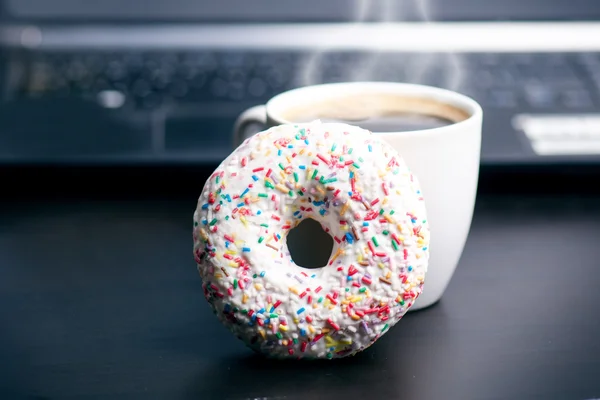Tasse Kaffee mit Donut vorhanden. im Hintergrund ist der Computer. Kaffeeschaum. Essen, Trinken und Technologie-Konzept. — Stockfoto