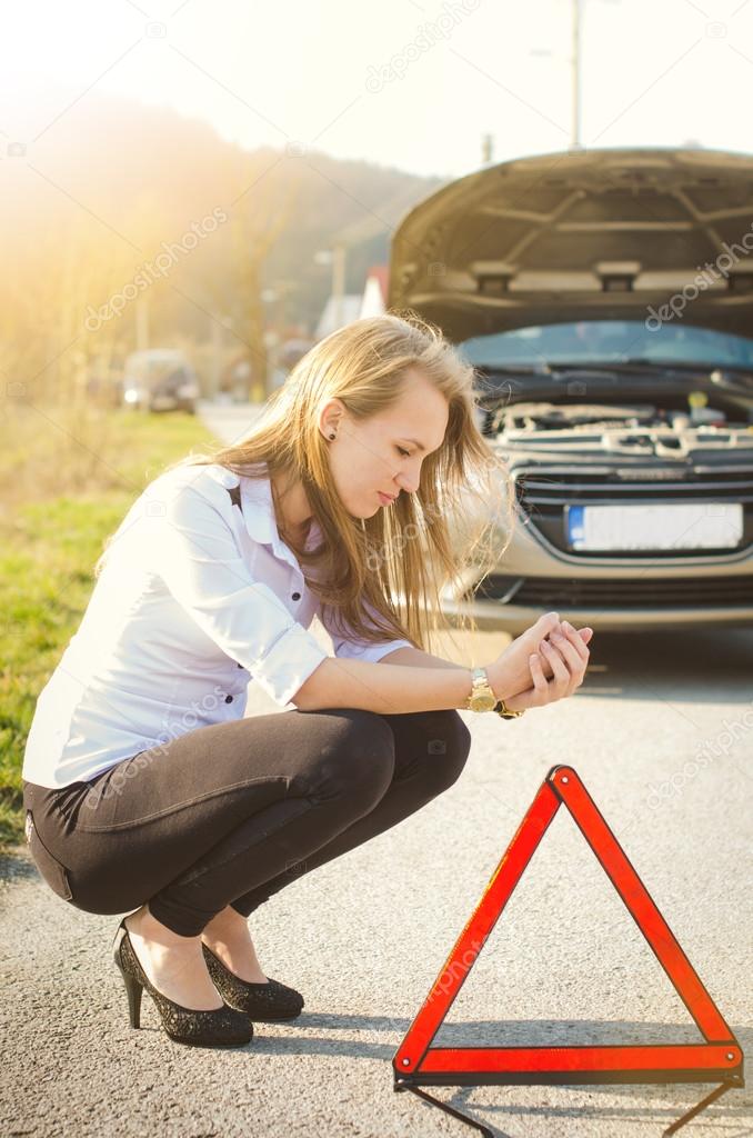 Woman crouching on the road next to a red warning triangle. Sad person. Damaged car. Natural background. Car accident.