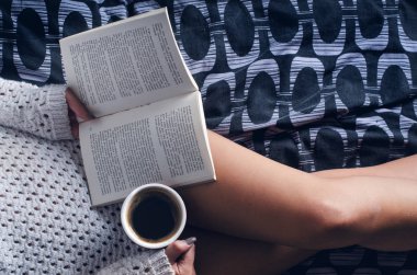Ayrıntı seksi genç kadın. Kadın bir fincan kahve tutmak ve bir kitap okuyun. Bir battaniye üzerinde oturuyor... Çıplak bacaklar. Ev atmosfer. 