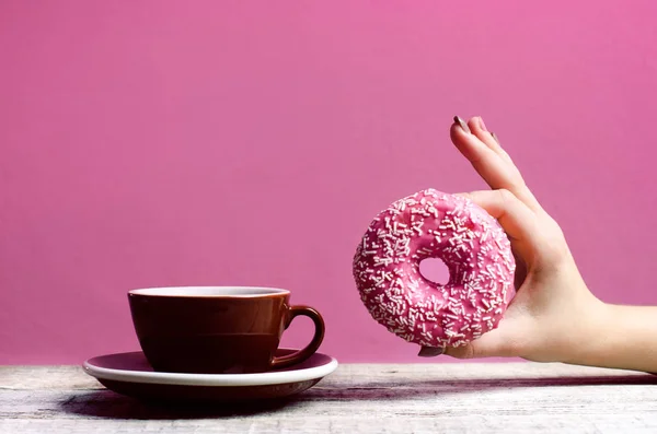 Hand hält bunten Donut mit Streusel auf einem Holztisch und rosa Hintergrund. Tasse Kaffee. zwei Arten von Donuts. Kuchen und Süßes. Lebensmitteldetails. Nahaufnahme. Pastellfarbe — Stockfoto