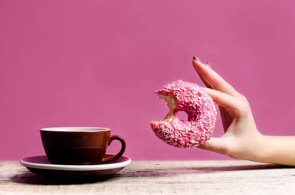 Frau Hand hält bunten Donut mit Streusel auf einem Holztisch und rosa Hintergrund. Tasse Kaffee. Konzept der Ernährung. Donut mit Biss fehlt aus nächster Nähe. Pastellfarbe — Stockfoto