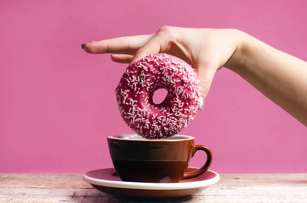 Frau Hand hält bunten Donut mit Streusel auf einem Holztisch und rosa Hintergrund. Tasse Kaffee. Konzept der Ernährung. Donut mit Biss fehlt aus nächster Nähe. Pastellfarbe — Stockfoto