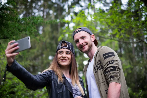 Pareja joven y moderna sosteniendo un teléfono móvil y riendo. Concepto de relaciones modernas. Primer plano de la gente hipster sentado en un bosque en el bosque . — Foto de Stock