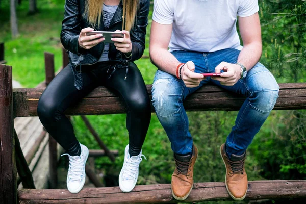 Pareja joven y moderna sosteniendo un teléfono móvil y riendo. Concepto de relaciones modernas. Primer plano de la gente hipster sentado en un bosque en el bosque . — Foto de Stock