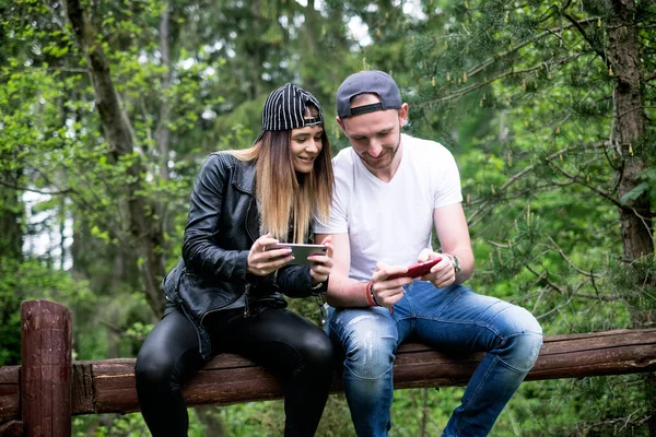 Pareja joven y moderna sosteniendo un teléfono móvil y riendo. Concepto de relaciones modernas. Primer plano de la gente hipster sentado en un bosque en el bosque . — Foto de Stock