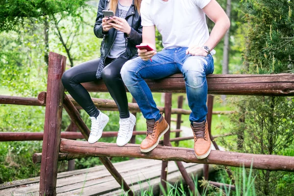Pareja joven y moderna sosteniendo un teléfono móvil y riendo. Concepto de relaciones modernas. Primer plano de la gente hipster sentado en un bosque en el bosque . — Foto de Stock