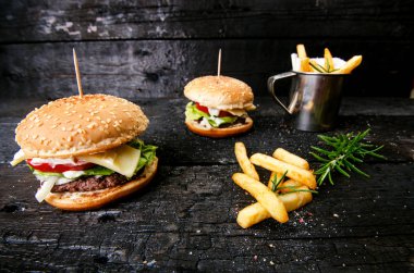 Yanmış, siyah ahşap bir masada patates kızartmalı hamburger. Fast food yemeği. Ev yapımı hamburger sığır eti, marul, domates, teneke, sos, peynir ve baharatlardan oluşur. Eski