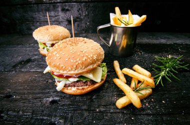 Yanmış, siyah ahşap bir masada patates kızartmalı hamburger. Fast food yemeği. Ev yapımı hamburger sığır eti, marul, domates, teneke, sos, peynir ve baharatlardan oluşur. Eski