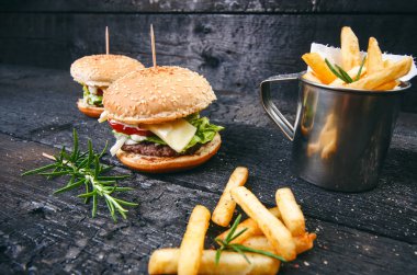 Yanmış, siyah ahşap bir masada patates kızartmalı hamburger. Fast food yemeği. Ev yapımı hamburger sığır eti, marul, domates, teneke, sos, peynir ve baharatlardan oluşur. Eski