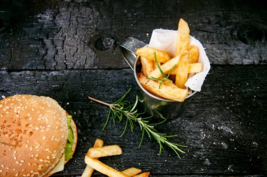 Yanmış, siyah ahşap bir masada patates kızartmalı hamburger. Fast food yemeği. Ev yapımı hamburger sığır eti, marul, domates, teneke, sos, peynir ve baharatlardan oluşur. Eski