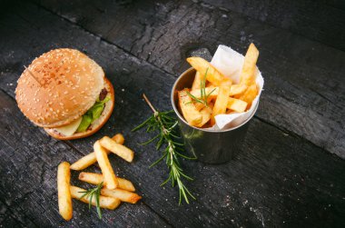 Yanmış, siyah ahşap bir masada patates kızartmalı hamburger. Fast food yemeği. Ev yapımı hamburger sığır eti, marul, domates, teneke, sos, peynir ve baharatlardan oluşur. Eski