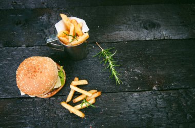 Yanmış, siyah ahşap bir masada patates kızartmalı hamburger. Fast food yemeği. Ev yapımı hamburger sığır eti, marul, domates, teneke, sos, peynir ve baharatlardan oluşur. Eski