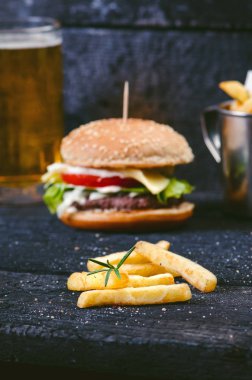 Yanmış, siyah ahşap bir masada patates kızartmalı hamburger. Fast food yemeği. Ev yapımı hamburger sığır eti, marul, domates, teneke, sos, peynir ve baharatlardan oluşur. Eski