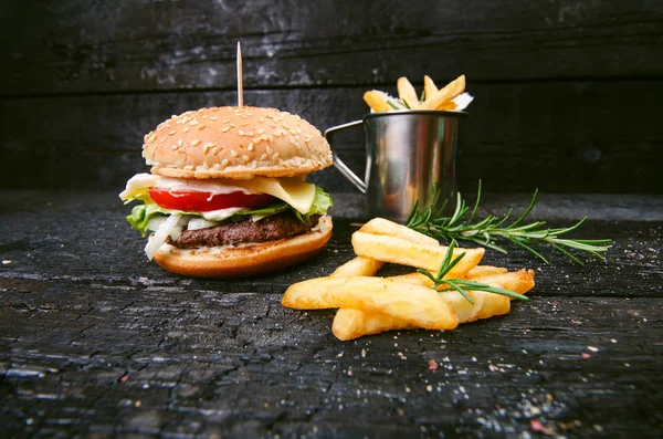 Yanmış, siyah ahşap bir masada patates kızartmalı hamburger. Fast food yemeği. Ev yapımı hamburger sığır eti, marul, domates, teneke, sos, peynir ve baharatlardan oluşur. Eski