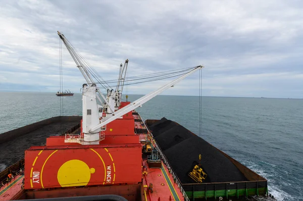 Loading coal from cargo barges onto a bulk vessel using ship cranes.