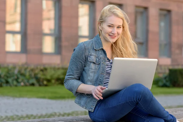Jovem estudante loira sentado em um banco e trabalhando em seu laptop — Fotografia de Stock