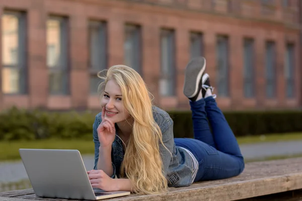 Jovem estudante loira deitado em um banco e trabalhando em seu laptop — Fotografia de Stock