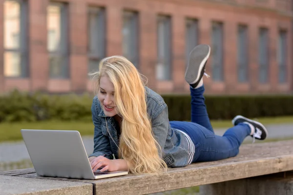 Jovem estudante loira deitado em um banco e trabalhando em seu laptop — Fotografia de Stock