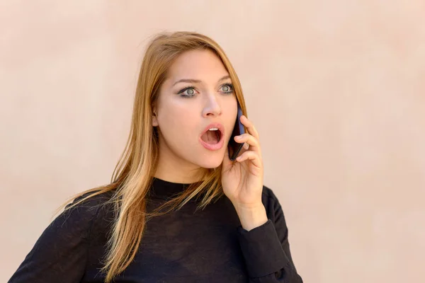 Attractive woman with a shocked expression chatting on her mobile phone staring ahead with mouth agape and wide eyes