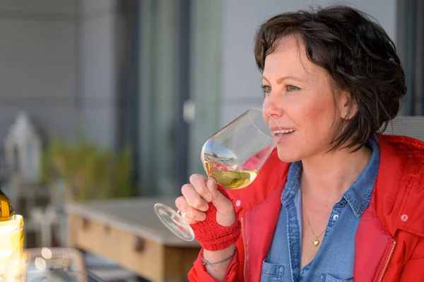 Trendy woman sipping a glass of white wine as she sits at an open air restaurant table in a close up profile view with thoughtful expression