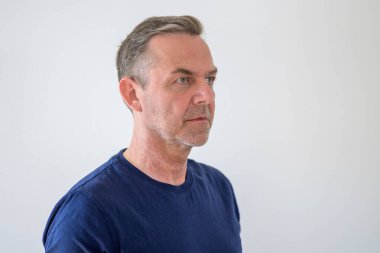 Kafasında ve omuzlarında beyaz bir portre olan mavi tişörtlü, traşsız, ciddi bir orta yaşlı adam çerçevenin sağına, boş kopya alanına bakıyor.