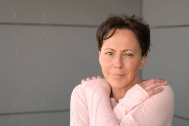 Çekici, orta yaşlı, Latin bir kadın, elleri omuzlarının üzerinde yatarken sessiz, düşünceli bir gülümsemeyle kameraya bakıyor.