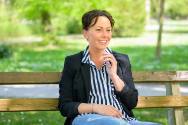 Şık bir kadın, bahar günü parkta keyif çatıyor bankta oturuyor ve elinde cep telefonu ve mutlu bir gülümsemeyle yan tarafa bakıyor.