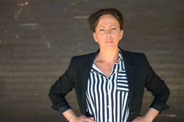 Özgüvenli İspanyol kadın yoğun bir ifadeyle kameraya dikkatlice bakıyor ve ellerini kalçasında gri bir dış duvara dayıyor.