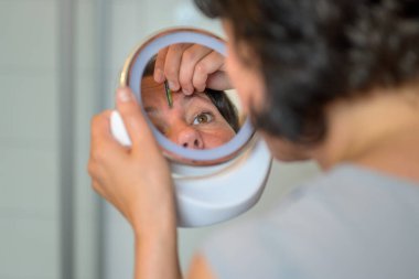 Omuz manzarası üzerinde küçük bir portatif ayna kullanarak kaşlarını yolan kadın yansımasına odaklanıyor.