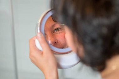 Omuz manzarası üzerinde küçük bir portatif ayna kullanarak kaşlarını yolan kadın yansımasına odaklanıyor.