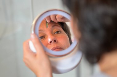 Bir kadın cımbızla kaşlarını yoluyor. Yakından yansımasına odaklanmış bir el aynası kullanıyor.