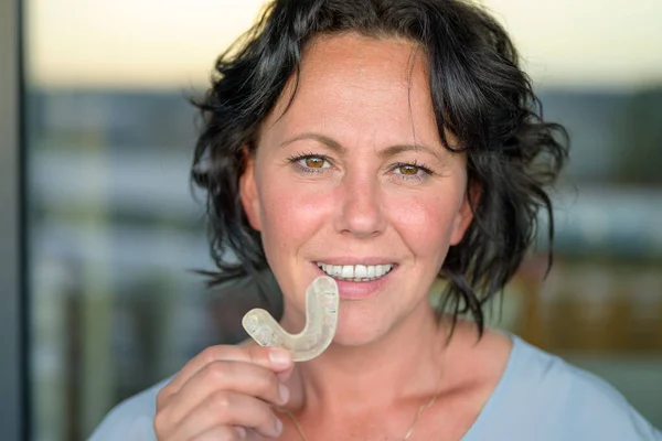 Attractive smiling woman suffering with bruxism holding a mouth guard in front of her face which she uses to protect her teeth from grinding