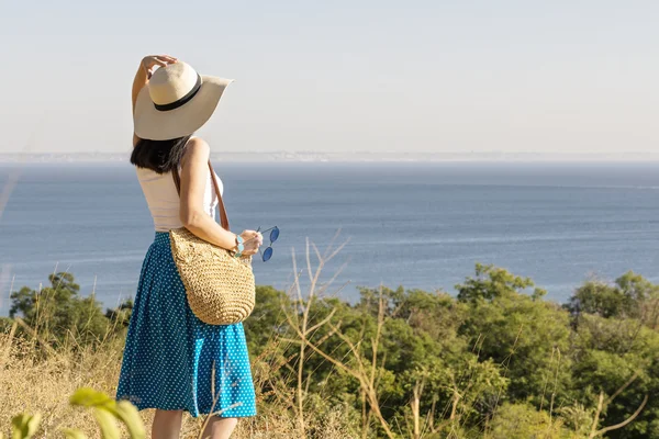 Young woman (brunette) in a blue skirt and hat looks at sea.