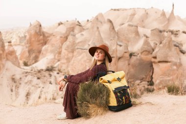 Çöl vadisinde kahverengi şapkalı, uzun elbiseli, boho tarzı sarı sırt çantalı gezgin bir kadın. Seyahat ve seyahat tutkusu konsepti. Kapadokya, Türkiye.