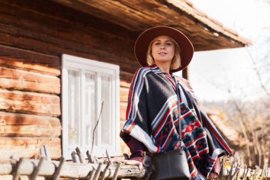 Kaliteli hippi gezgin kadın otantik boho şıklığında panço ve eski ahşap kırsal binaların yakınında şapka takıyor. Sonbaharda doğayı keşfeden kadın. Seyahat ve seyahat tutkusu kavramı..