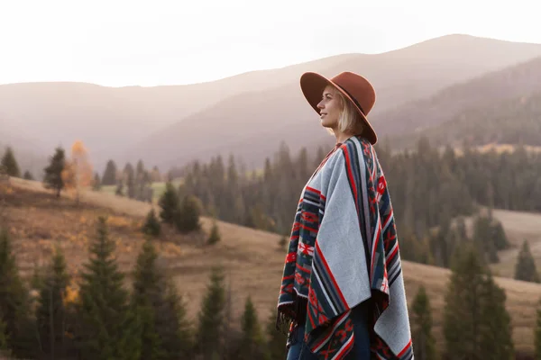 秋の風景の中で本格的なブーブーシックなスタイルのポンチョと帽子を身に着けているスタイリッシュなヒップスター旅行女性 幸せな若い女性は日当たりの良い山を探索 旅と放浪の概念 — ストック写真