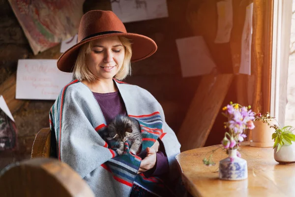 Pançolu ve şapkalı küçük tekir kedili kadın kırsal kabin odasındaki pencerenin yanında oturuyor. Yavaş yaşam ve evcil hayvan bakım konsepti.