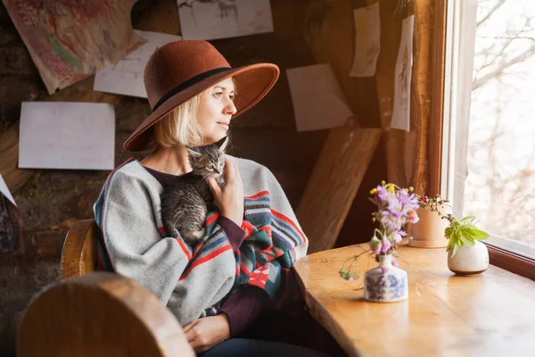 Pançolu ve şapkalı küçük tekir kedili kadın kırsal kabin odasındaki pencerenin yanında oturuyor. Yavaş yaşam ve evcil hayvan bakım konsepti.