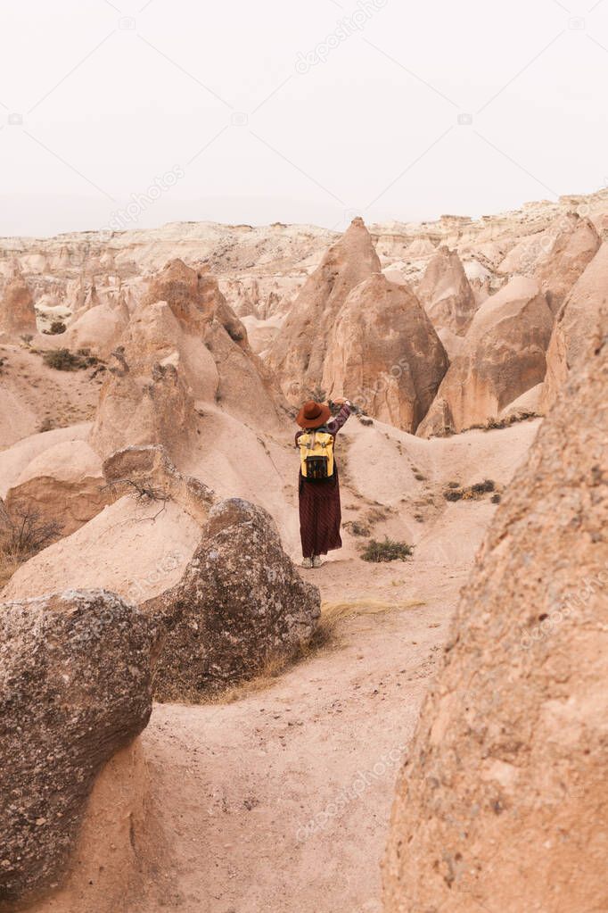 Travelling woman wearing brown hat and long dress with yeloow backpack in desert valley and rocks. Travel and wanderlust concept. Cappadocia, Turkey.