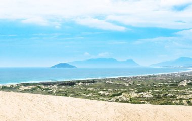 Plaj, kum, yeşil bitki örtüsü, mavi okyanus ve arka planda bazı dağlar