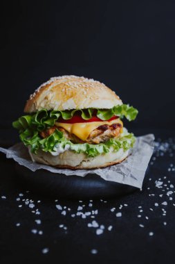 İştah açıcı taze ev yapımı burger, tavuk pirzolası, marul, domates, peynir ve sos. Siyah arka planda, tahta bir tahtanın üzerinde.